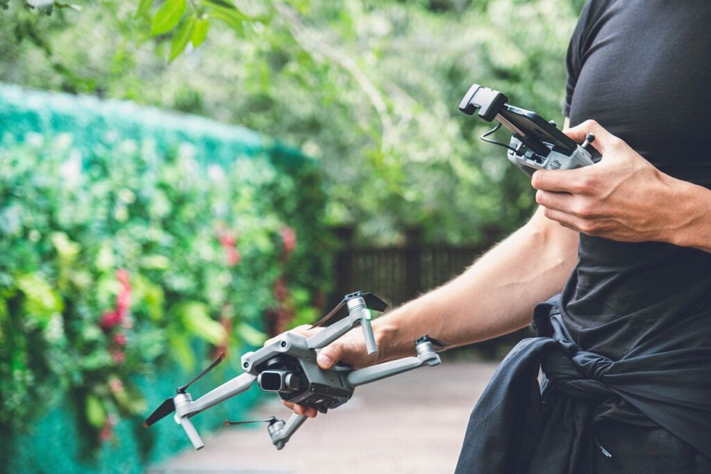 A person in a black shirt and black pants holding a gray drone and a black controller outside the house