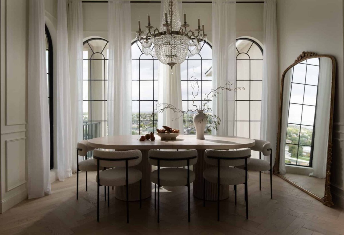 Dining room with a high ceiling, long, white draperies, classy chandelier and a huge mirror resting on one of the side walls