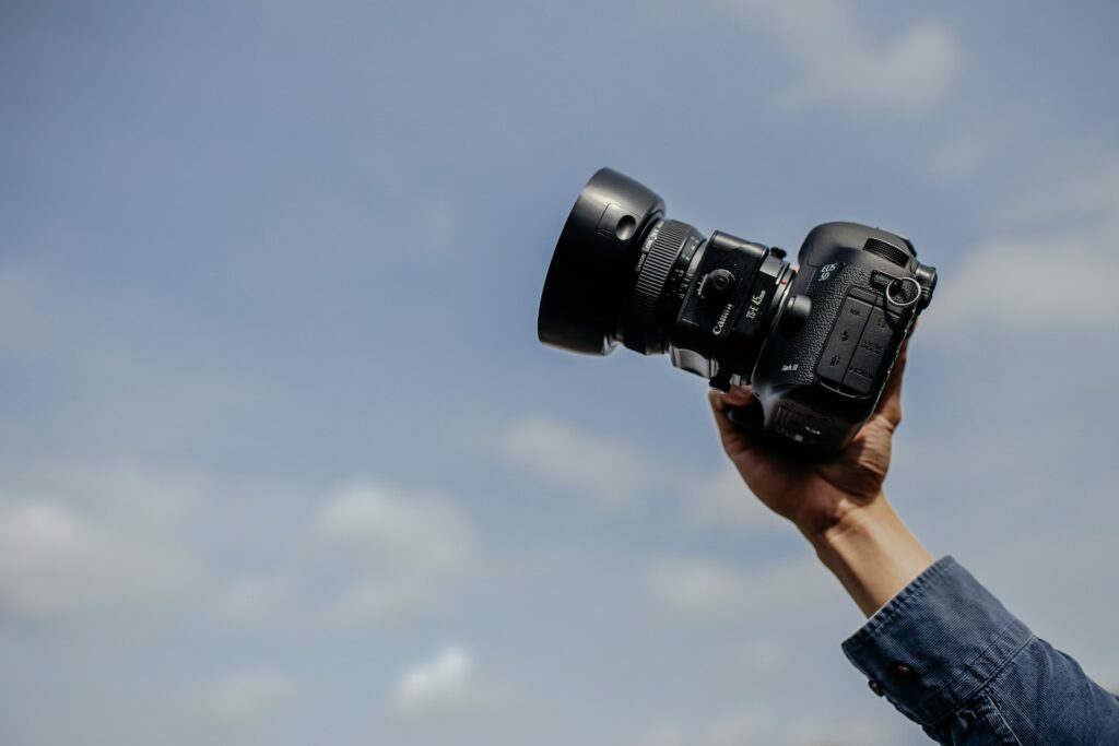A photographer wearing a denim long sleeves top while pointing a black SLR camera in a clear blue sky