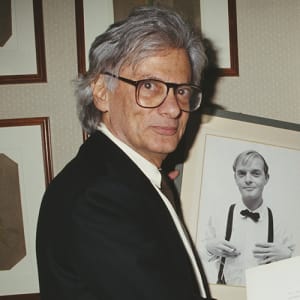 Richard Avedon wearing a black suit with a black tie near framed photographs on a cream-colored wall