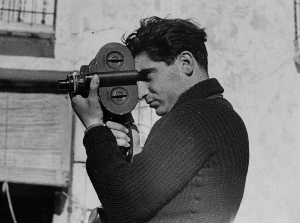 A black and white image of Robert Capa wearing a long sleeves knitted shirt while holding a camera