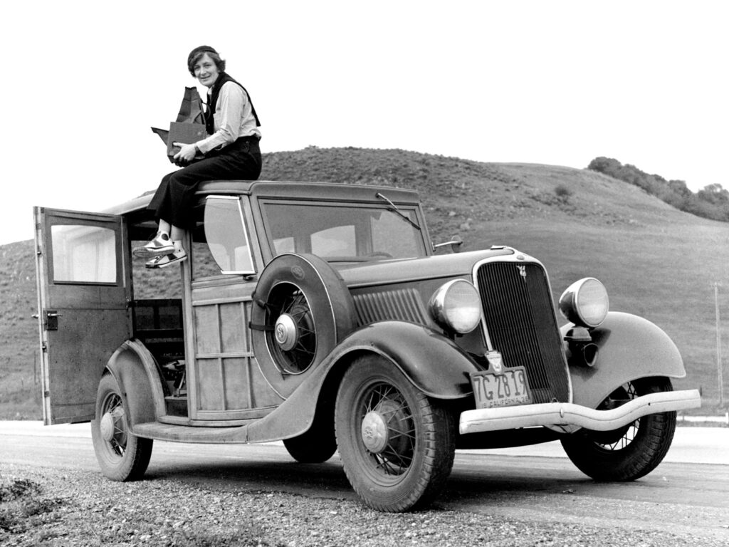 A black and white image of Dorothea Lange sitting on top of a car