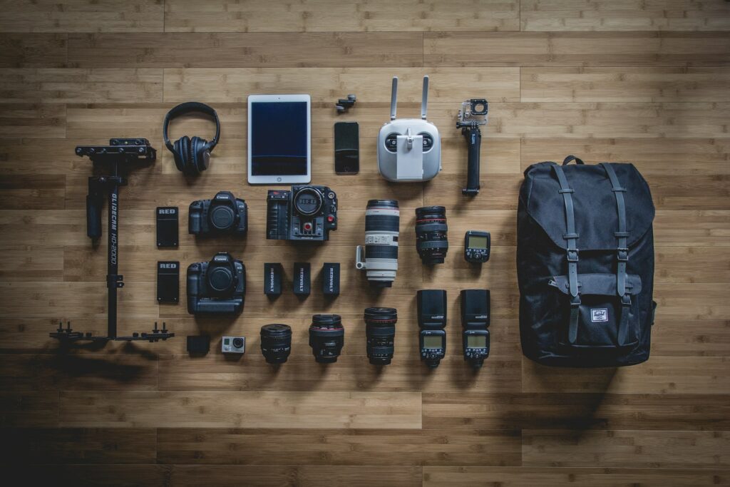 Three black DSLRs with black and white camera lenses, a black tripod, a white smart tablet, a black backpack, and other camera equipment on top of a brown wooden table