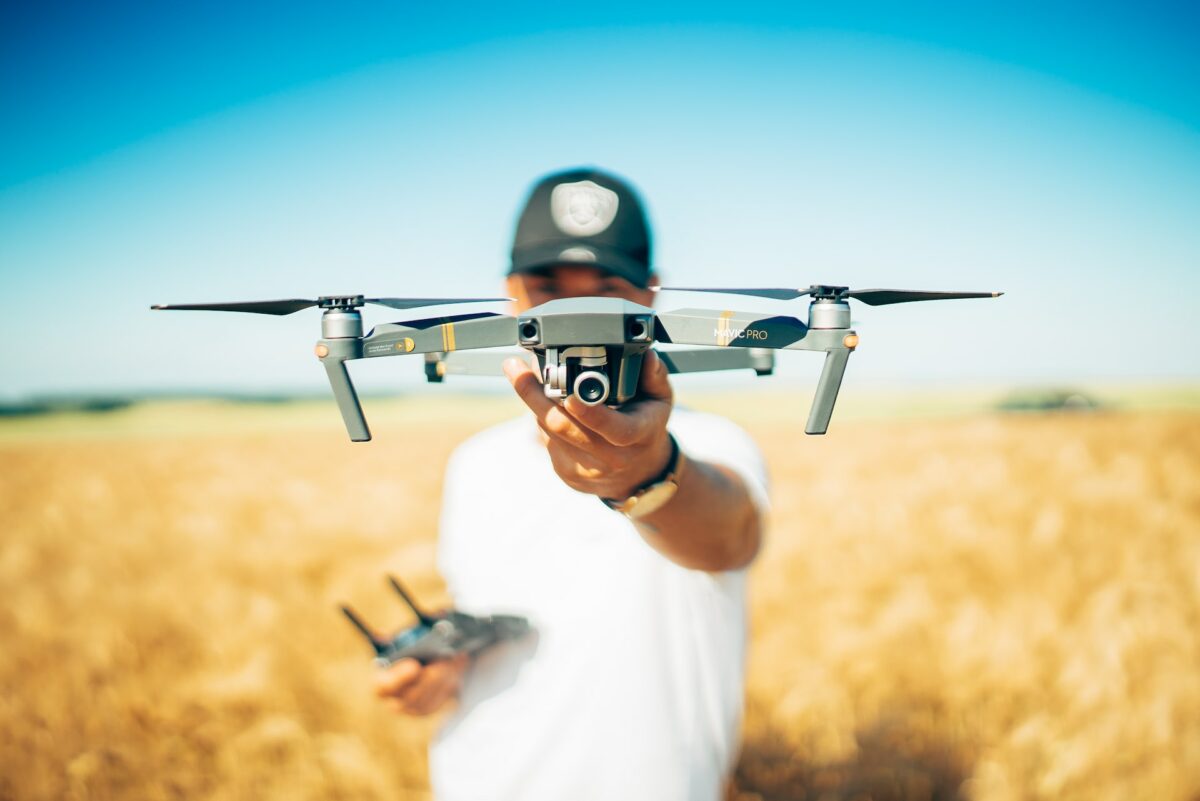 A man wearing a white shirt and black cap is holding a black drone on his left hand