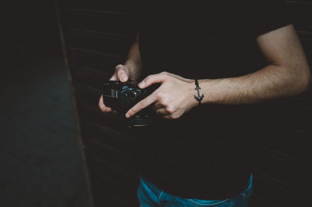 A person wearing a black shirt and blue jeans holding a black DSLR camera near a wooden wall