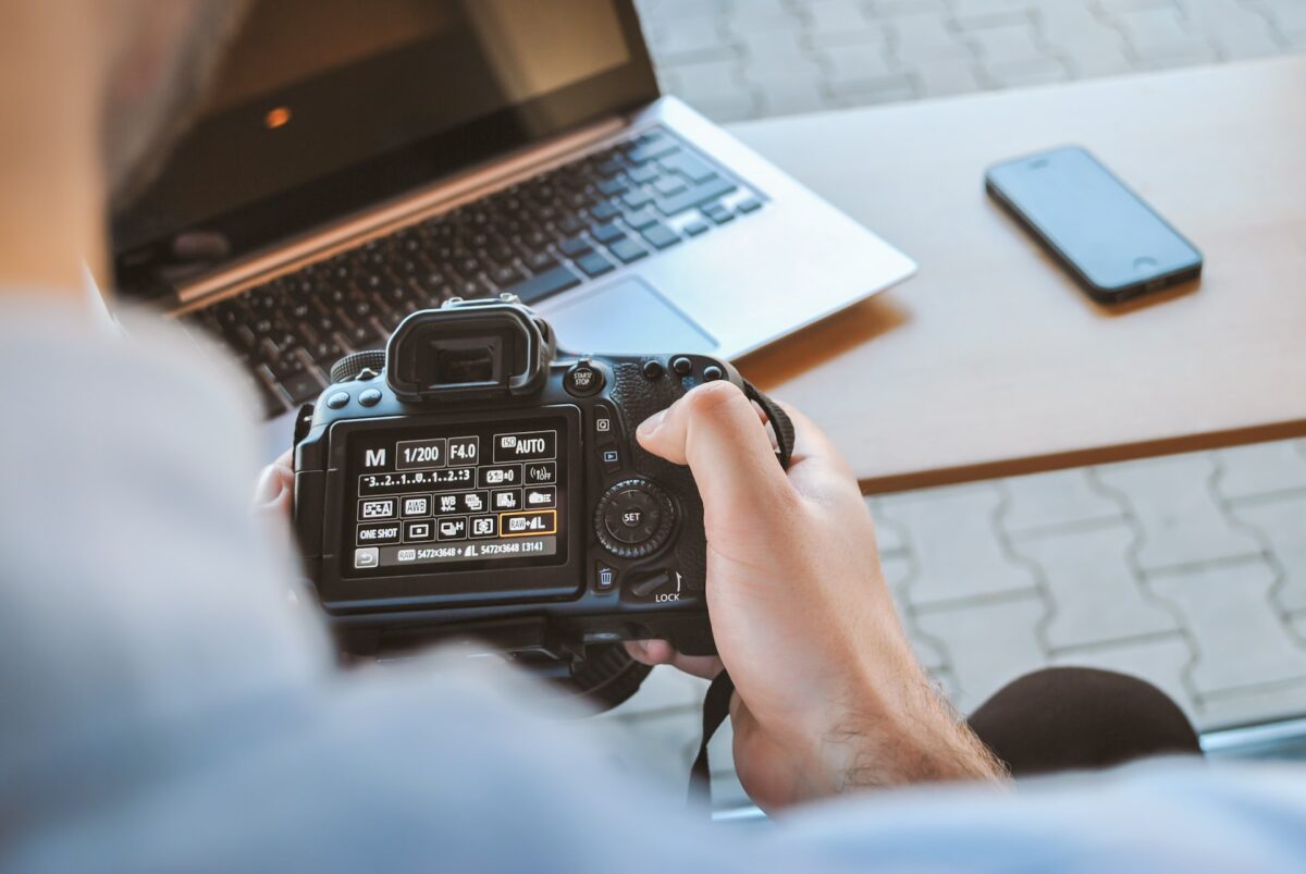 A person holding a black DSLR camera near a silver laptop and smartphone on top of a brown wooden table